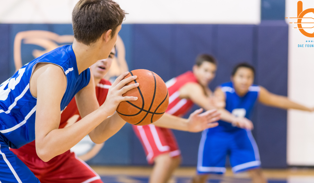 A young basketball player in a blue sleeveless jersey holding a basketball with two hands, looking down the court. In the blurred background, other youth players in red and blue jerseys move in defensive stances on an indoor gymnasium court, with an orange DAE Foundation logo in the top-right corner.